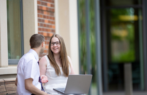 Man and woman sitting on a bench talking. The woman is holding a laptop, smiling, talking with the man to her left.