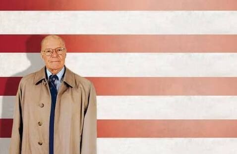 Old man in a trench coat standing in front of red and white striped background.