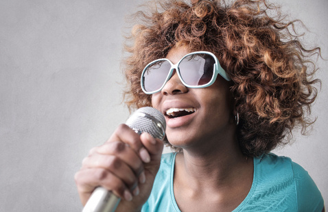 Close-up of a young woman with curly hair and sunglasses singing passionately into a microphone.