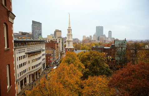 White church steeple rising above orange autumn trees amid downtown buildings and a street with pedestrians.