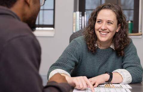 A woman smiles while discussing a scored worksheet with a student seated across the table, pointing to a specific section of the page.