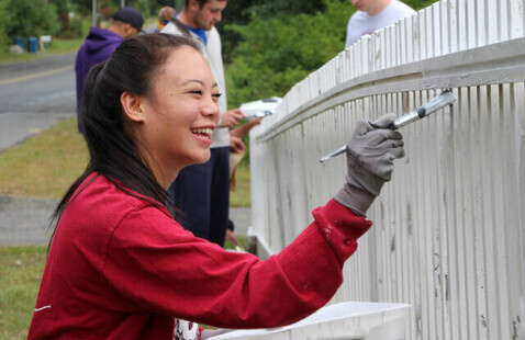 Woman smiling while painting a white picket fence, wearing a UMass shirt and gloves, volunteers in background