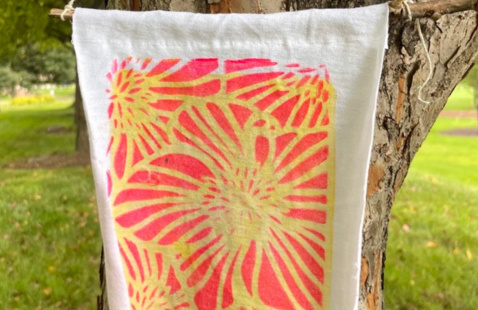 A white fabric banner hanging from a tree outdoors, featuring a bright pink and yellow abstract floral/sunburst pattern. The banner is photographed against a green park background with grass and trees.
