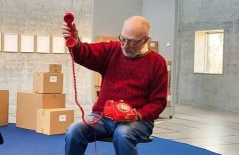 Headshot of Fluxus artist Ken Friedman. He is wearing a red shirt and holding a red telephone.