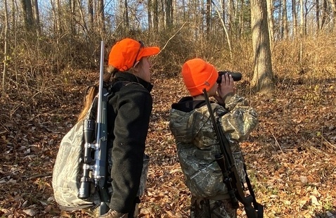 Two people in orange caps and camouflage carrying rifles, one looking through binoculars in a leaf-strewn deciduous woods.