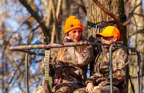 Adult woman and child in camouflage and orange caps on an elevated tree stand in leafless woods.