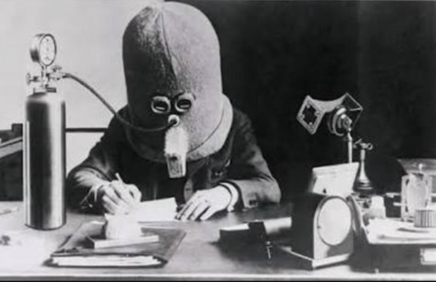 Man at desk wearing large hood with breathing tube to a gas cylinder, writing while surrounded by office instruments