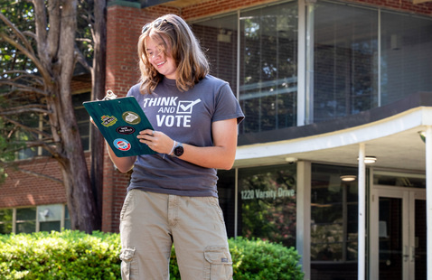 A student wearing a “Think and Vote” T-shirt holds a  hold a clipboard covered with civic engagement stickers while standing outside a campus building .