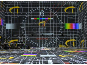 Room covered by projected test pattern: grid, color bars, large 6 and CREATIVE logo on walls and floor; AV gear left