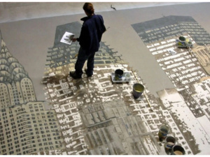 Artist standing on large floor mural of skyscrapers holding reference sheet, surrounded by paint cans and buckets