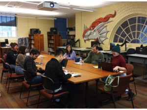 Group of people seated around a table with laptops and notebooks in a room with a dragon mural and arched window