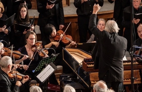 A conductor gestures in front of a group of string players on stage, who watch him intently 