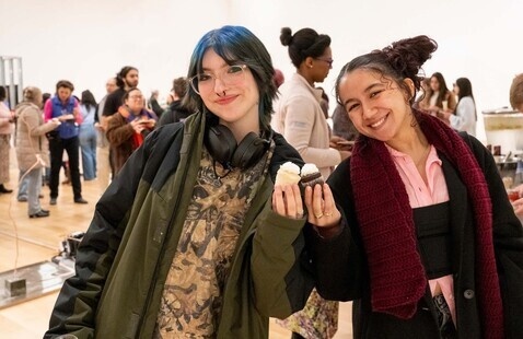 Two students smiling and holding cupcakes at an event with people in the background. Photo: Melissa Ostrow. 