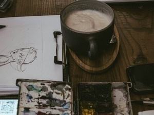 Workspace closeup: Drawing pad and paints next to a mug.