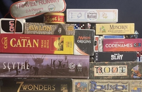 A wall of board games displaying games such as 7 Wonders, Dominion, Scythe, Root, Catan, Magic the Gathering, Here to Slay, Codenames, Avalon, Uno, Spot It!, Travel Rummy, Command of Nature, Munchkin, standard decks of cards, and a variety of dice. 