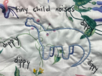 A hand-embroidered white fabric panel with colorful floral and abstract designs. In the center is an outlined dinosaur, surrounded by handwritten words including “tiny child noises" and “dippy,” with small flowers, leaves, and decorative shapes.