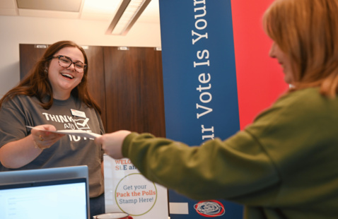 A student volunteer smiles while handing materials to another person at a Pack the Polls voter engagement event.