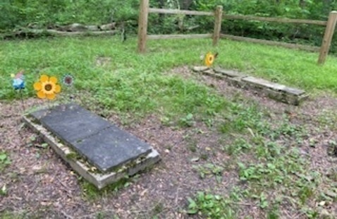two gravestones on concrete pads with grass around, a wooden fence and woods beyond. Old, weathered gray rock 