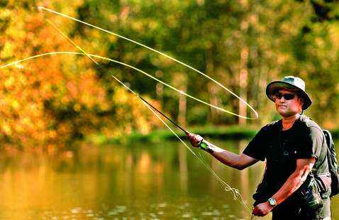 Man in sun hat and sunglasses fly-casting a rod at a tree-lined lake with autumn foliage.