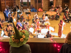A balcony view photograph of a previous Monsters Ball performance. The audience is in the foreground and the orchestra is performing on the floor in the background. Many people are wearing costumes.