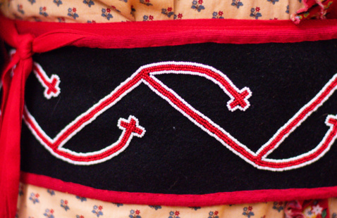 A close-up of a colorful sash with intricate patterns. The design features red, white, and black embroidery with a zigzag pattern and cross motifs. The sash is worn over a garment with a small floral pattern.