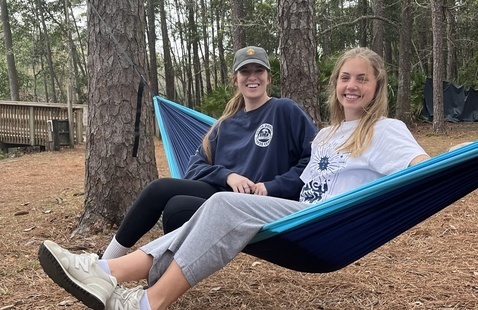 Two women sitting on a blue hammock surrounded by trees