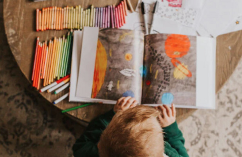 Overhead view: young child at round wooden table with open illustrated book and colored pencils