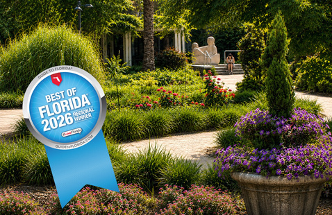 Landscape view of the UNF Botanical Garden featuring a winding garden path, flowering beds with pink and purple blooms, a large decorative planter, manicured evergreen shrubs, and a sculpture in the background. A Best of Florida 2026 Regional Winner ribbon graphic is displayed in the lower left corner.