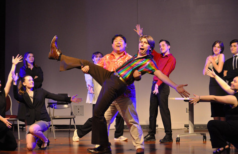 Students on a stage performing a song ordance. One student is holding another student whose arms are outstretched in a "ta-da" fashion.