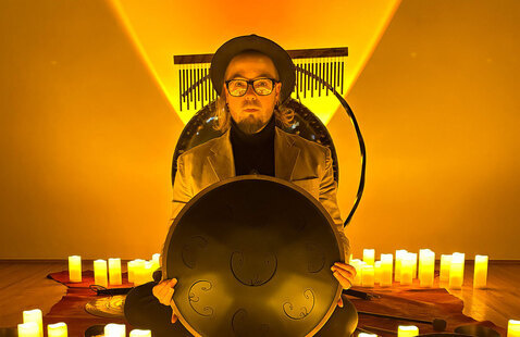 Harold Taddy holding a gong, sitting on a stage surrounded by candles.