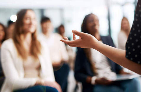 Instructor's outstretched hand gesturing to seated adult audience in a meeting room.