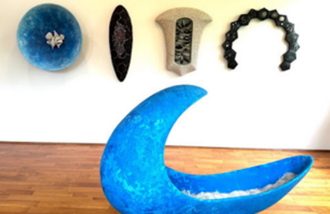 Large blue crescent sculpture with four wall-mounted items above: blue disk, black oval, organic beige T-shape, black arch.