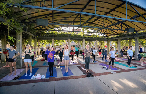 group of 25 or so people on yoga mats on the stage at the uptown community park, doing yoga.