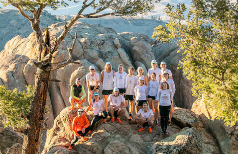About 15 runners in matching shirts posing on rocky overlook among juniper trees with distant mountain range