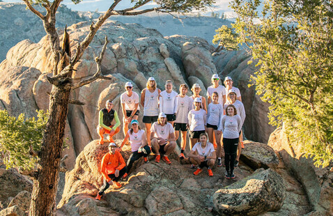 Group of people wearing matching white shirts posing on rocky outcrop among trees with distant mountains