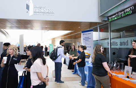 Students and recruiters interact at a career fair inside a bright, modern university building. Booths line the hallway beneath signage for the College of Business Administration, with attendees networking, shaking hands, and exchanging information in a professional campus setting.