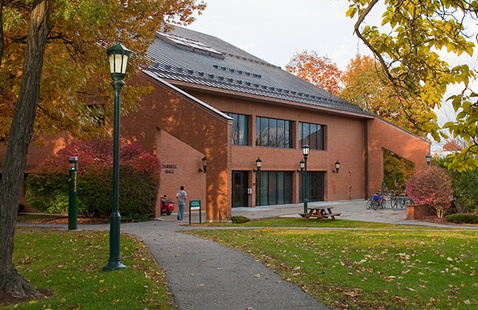 Brick campus building with students at entrance, lamp posts, picnic bench and fall foliage on a paved path