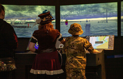 Two people wearing Halloween costumes stand in front of ship simulator screens.