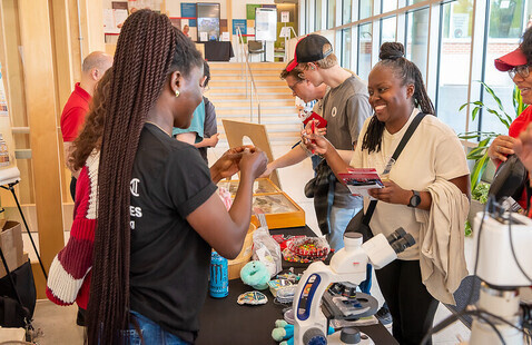 Several people interacting at a table that includes a microscope and plant samples.