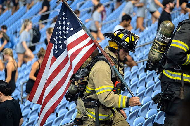 Firefighter in turnout gear carrying American flag up stadium steps with blue seats and spectators