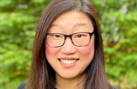 Smiling woman with glasses, wearing black top and statement necklace, blurred green foliage background.