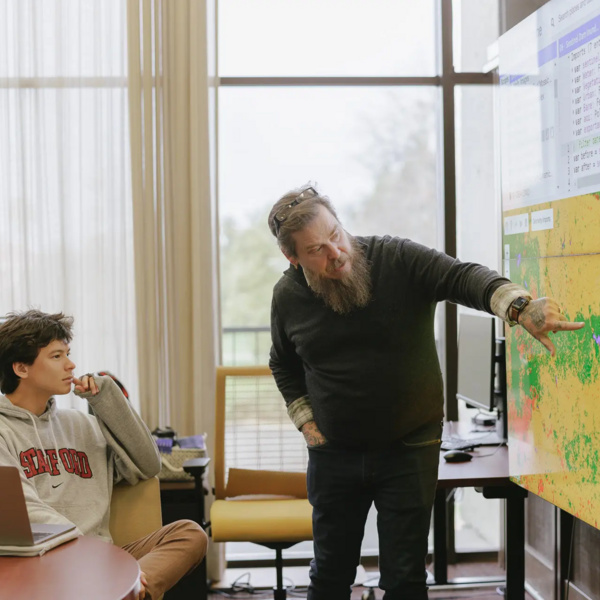 Instructor pointing at large map on screen as seated student wearing STANFORD sweatshirt watches in conference room