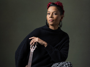 Musician Rhiannon Giddens sits barefoot against a dark backdrop wearing a black sweater and checkered skirt, restfully holding a banjo upright on the floor.