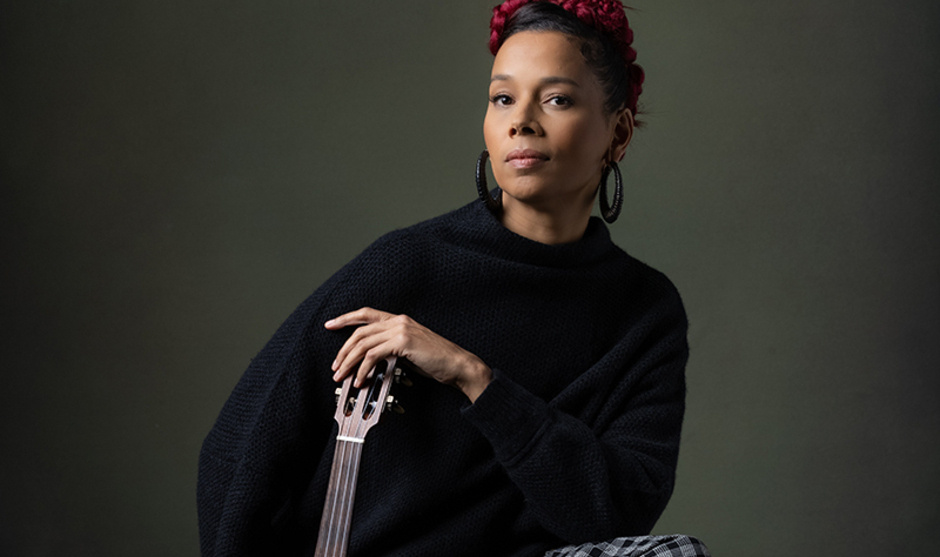 Musician Rhiannon Giddens sits barefoot against a dark backdrop wearing a black sweater and checkered skirt, restfully holding a banjo upright on the floor.