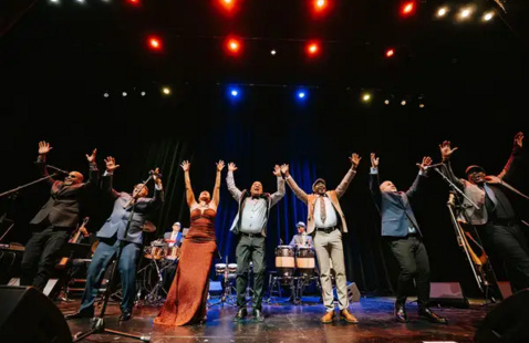 Eight performers stand on stage raising arms. In the center is a vocalist in red gown. Behind them are percussionists and colored stage lights.