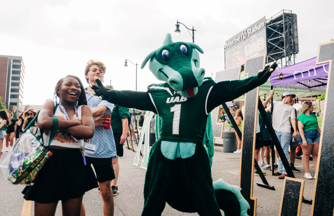 Blaze is standing beside two smiling students outside of Protective Stadium at a UAB Football Tailgate. 