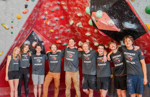 Ten climbers in matching WESTERN COLORADO shirts standing arm-in-arm in front of a red indoor bouldering wall