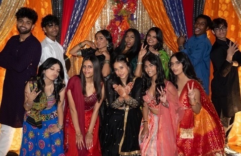 Group of young adults in colorful South Asian attire making hand signs and posing before orange-gold draped backdrop