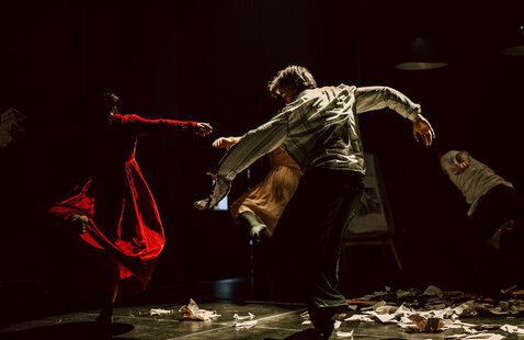 Photograph captures a contemporary dance performance on a dark stage, with central dancer extending arms amid motion and dramatic lighting. Multiple performers surround dancer, including figure in flowing red costume, while scattered papers and overhead spotlights emphasize theatrical atmosphere and movement.