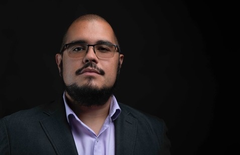 Mauricio Castro with glasses and beard wearing dark suit jacket and lavender shirt, facing camera against a black background.